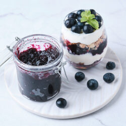 blueberry jam in a glass jar alongside parfait made with blueberry jam and yogurt.