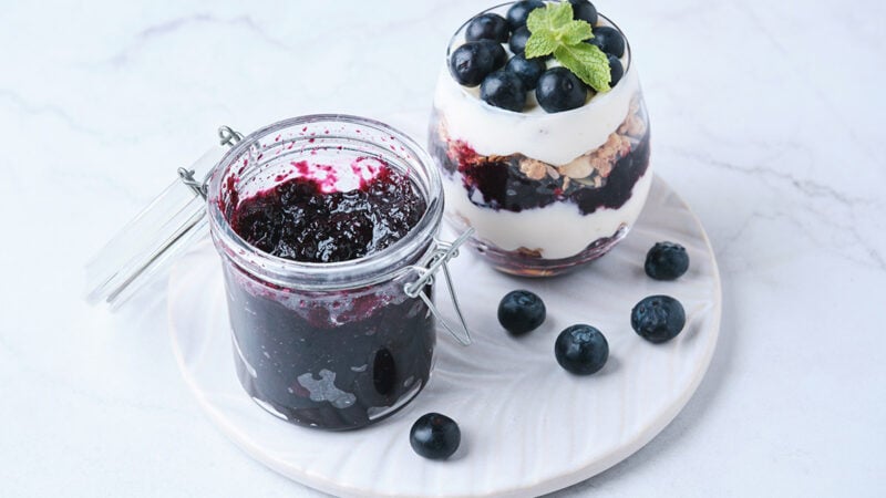 blueberry jam in a glass jar alongside parfait made with blueberry jam and yogurt.