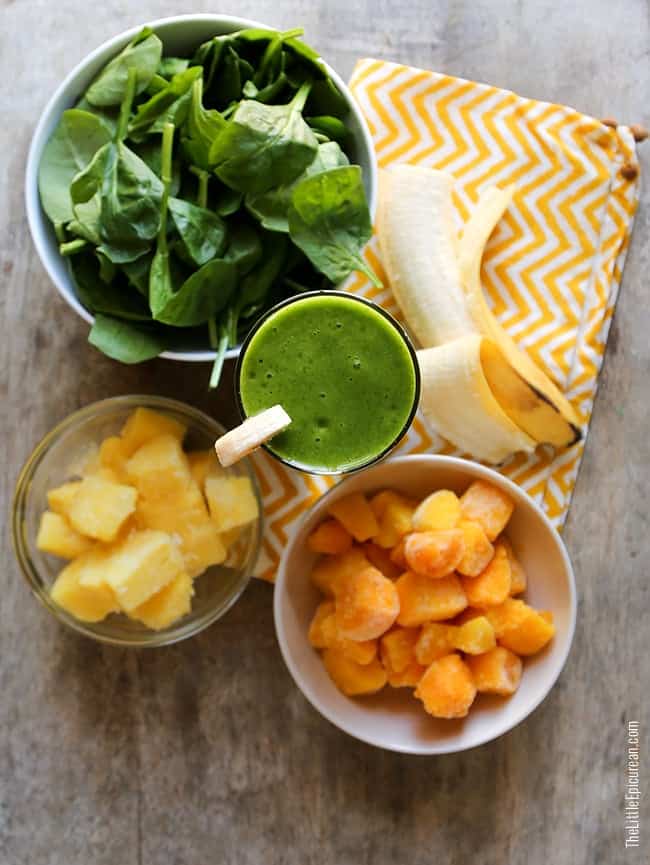 overhead view of a glass of green smoothie surrounded by frozen mango chunks, fresh banana, and spinach leaves. 