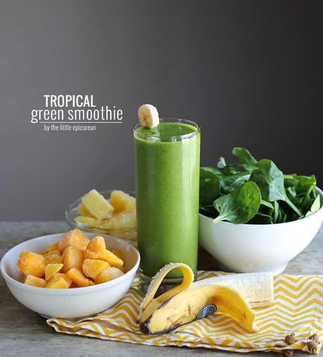 a glass of green smoothie surrounded by frozen mango chunks, fresh banana, and spinach leaves.