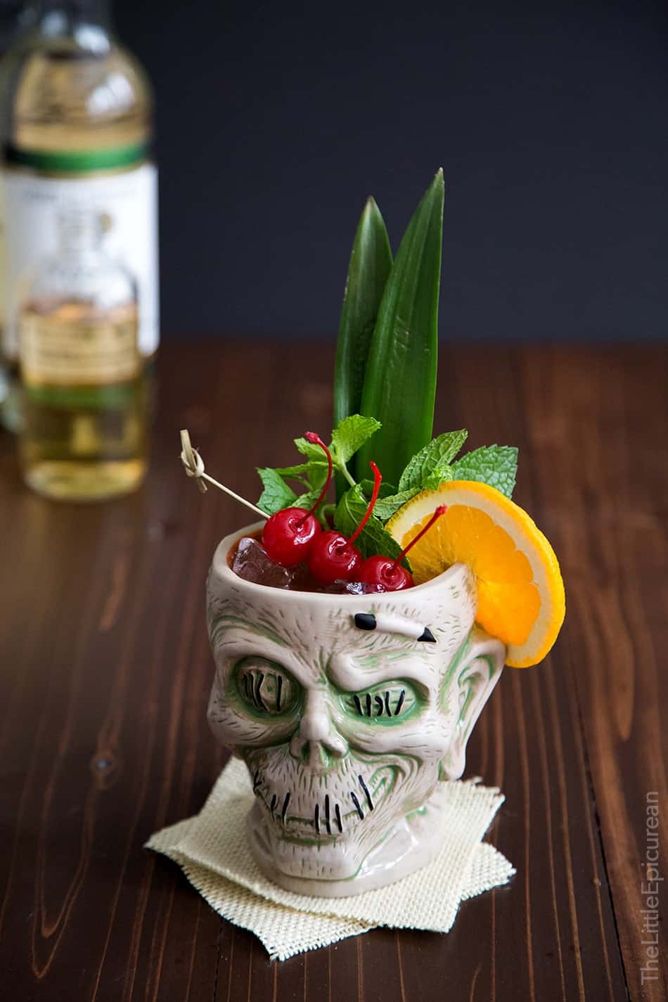 tiki cocktail garnished with cherries, mint, and orange slice served in a zombie mug glass.