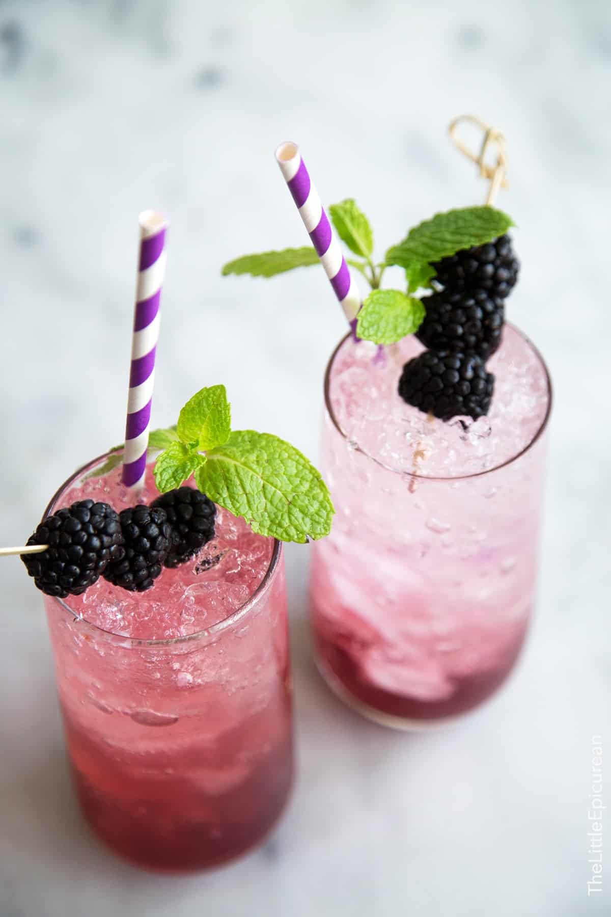 two servings for sparkling blackberry shrub mocktails garnished with fresh blackberries, mint, and purple straw.