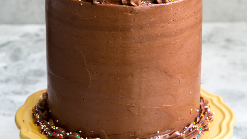 layered yellow cake coated in chocolate frosted and topped with sprinkles displayed on a yellow cake stand