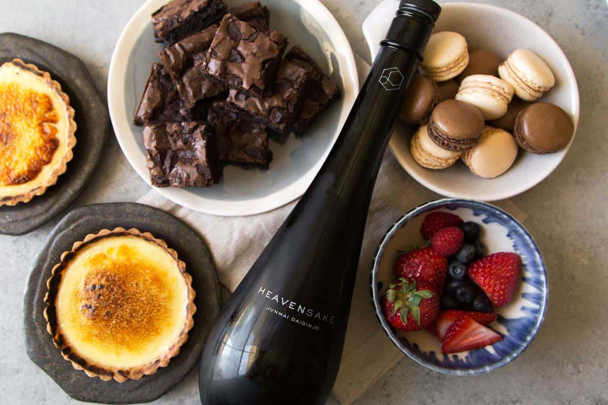 bottle of sake surrounded by creme brulee tart, brownies, macarons, and fresh berries.