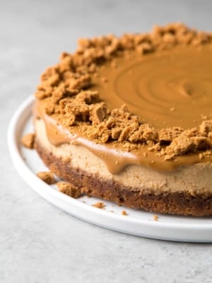 biscoff cheesecake topped with melted cookie butter and garnished with crushed biscoff cookies on white serving platter.
