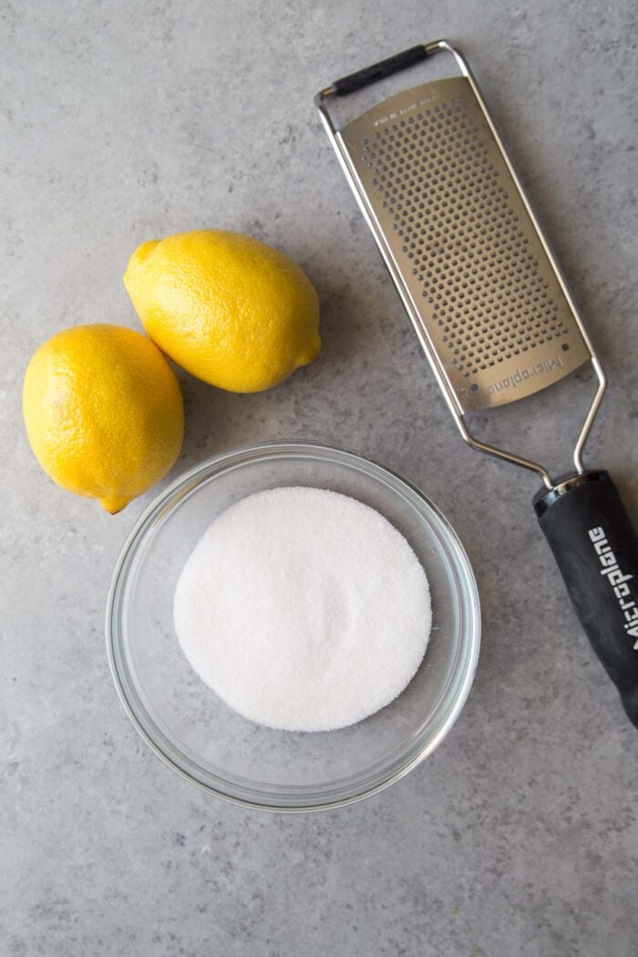 two fresh lemons, a bowl of granulated sugar, and a microplane zester.