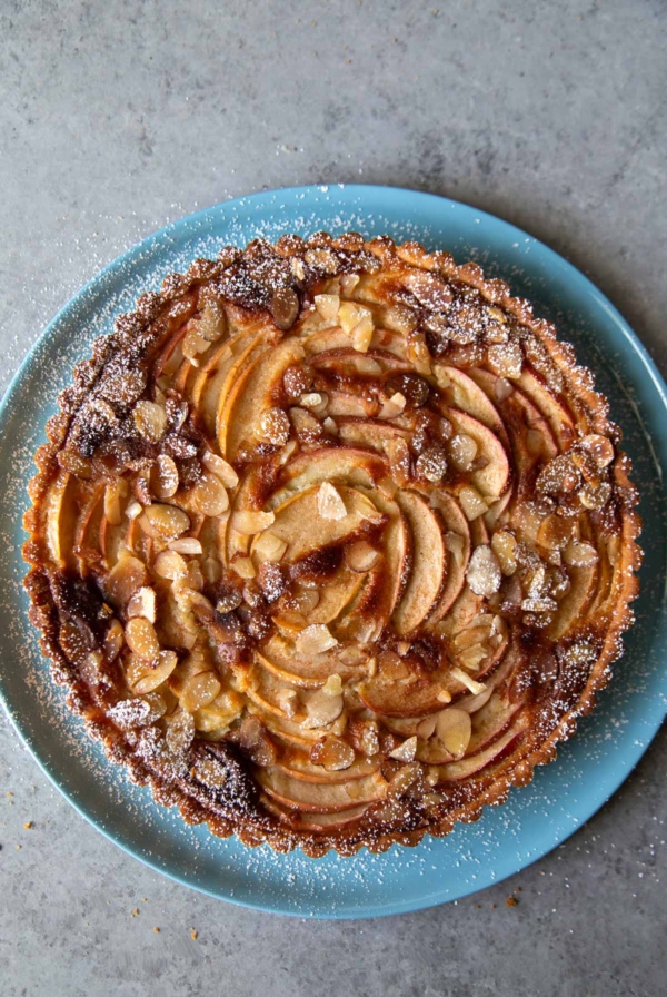 overhead of apple frangipane tart dusted with powdered sugar on blue serving plate.