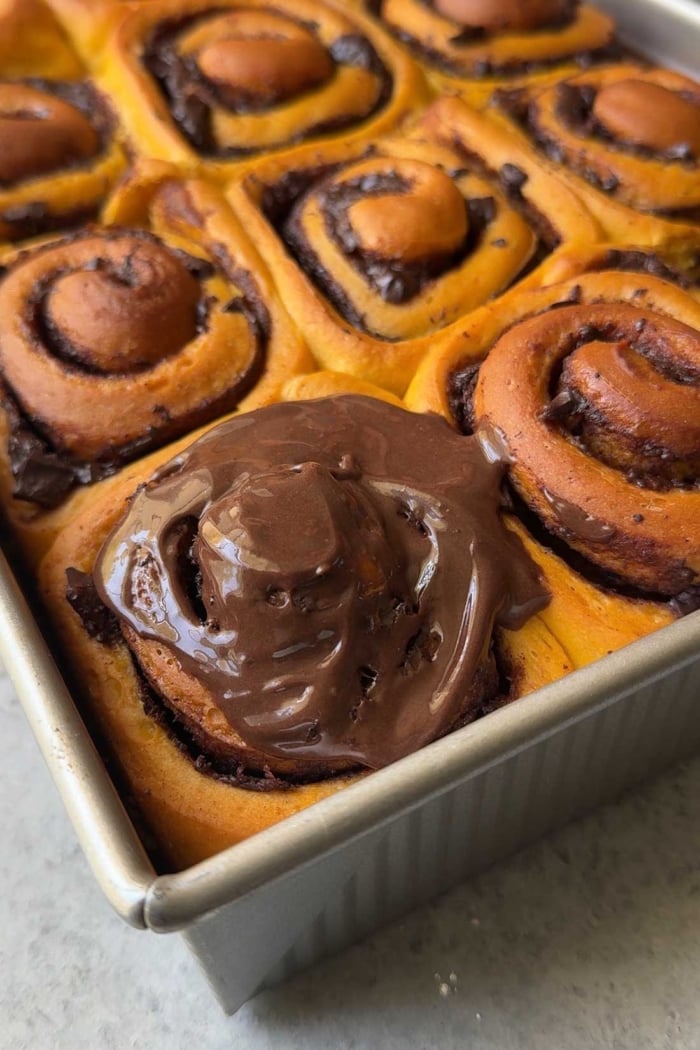 chocolate pumpkin cinnamon rolls in baking pan with one roll frosted.